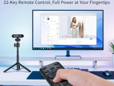 EMEET C60E Dual-Camera 4K Webcam on a small tripod beside a monitor, a hand holding its 22-key remote in the foreground
